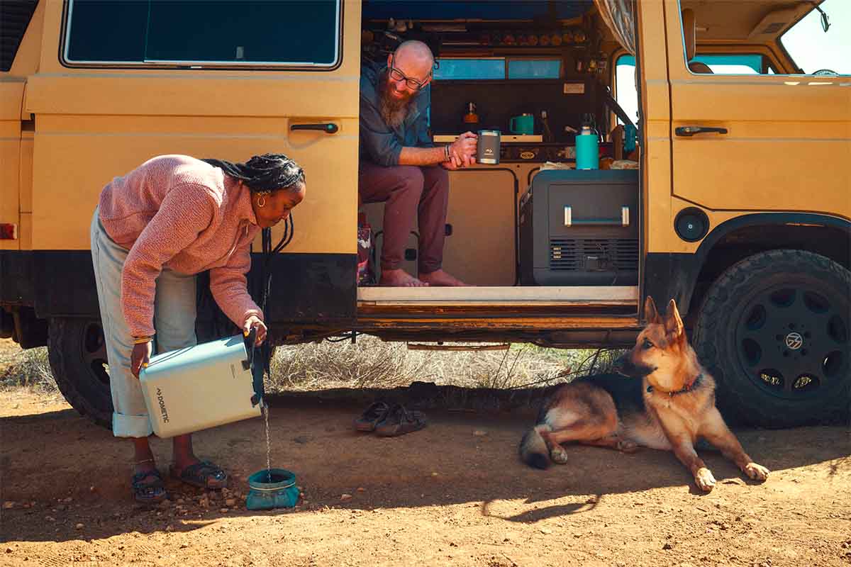 un minivan équipé pour l'overlanding et un couple avec un chien. Il y a un grand réservoir d'eau dometic pour le chien